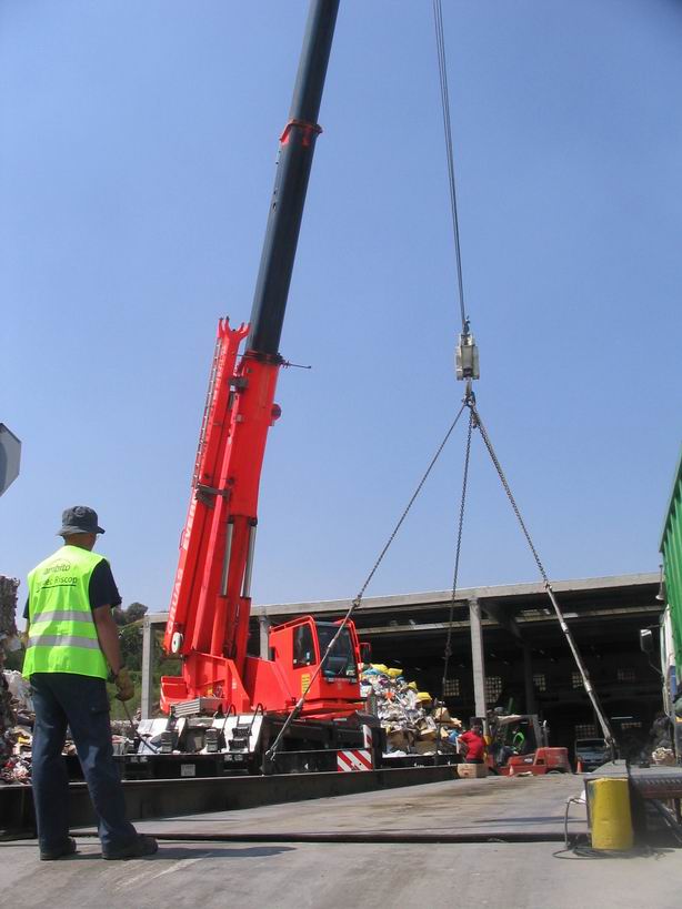 Técnico de TecniPés con chaleco fluorescente trabajando sobre una báscula puente para camiones enrasada en el suelo, con grúa de elevación al fondo.