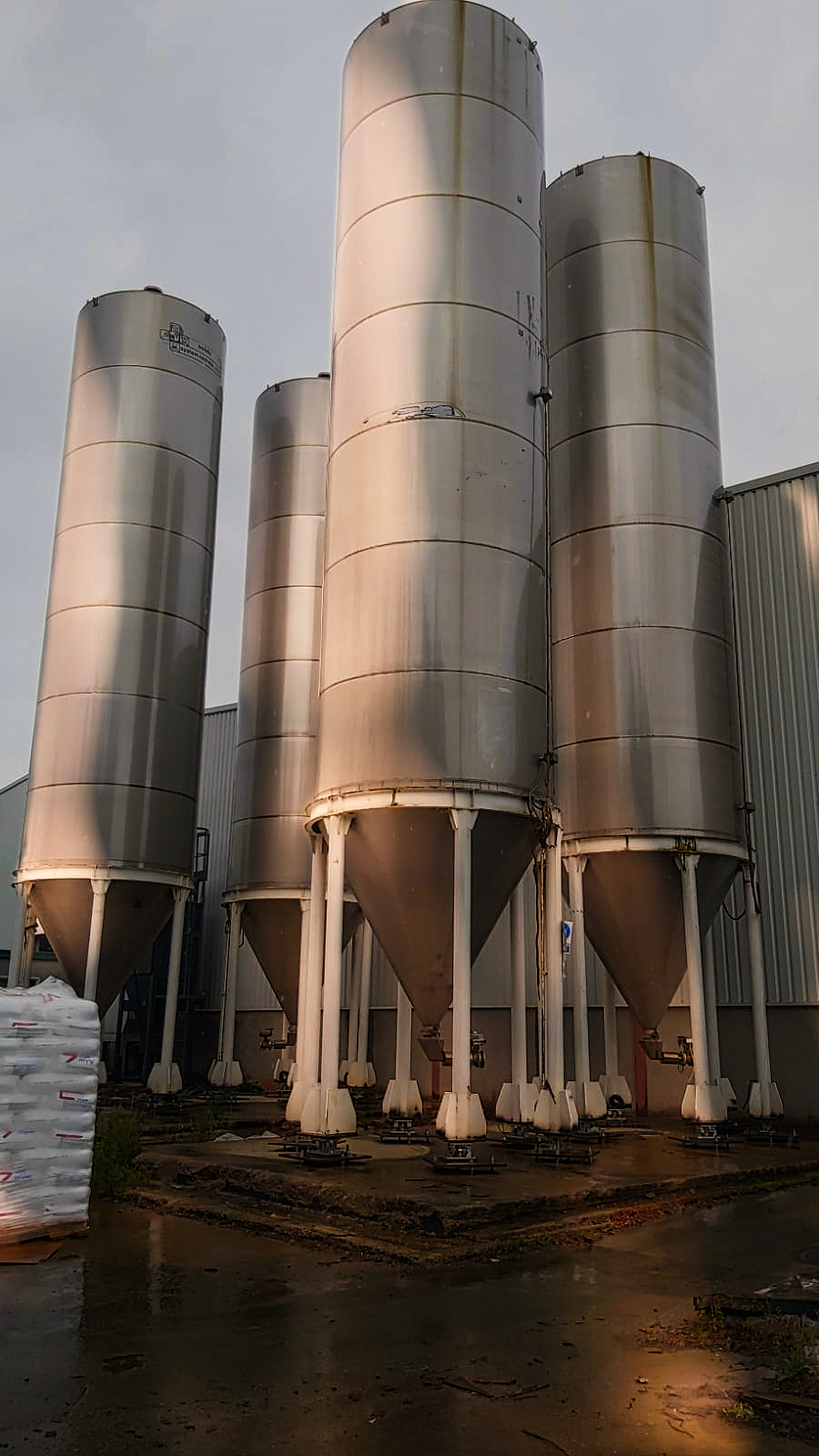 Seis silos verticales de acero inoxidable con pies cónicos blancos para pesaje de producto a granel en planta industrial.
